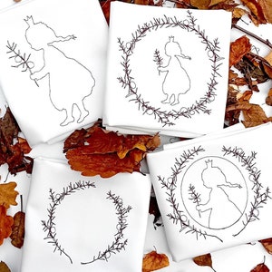 Puede incluir: Cuatro cuadrados de tela blanca con diseños bordados. Dos cuadrados presentan a una niña con una corona y sosteniendo una rama. Los otros dos cuadrados presentan una corona de ramas con bayas.