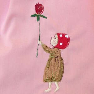 Könnte beinhalten: Ein rosa Stoff mit einer gestickten Darstellung eines Mädchens, das eine rote Blume hält. Das Mädchen trägt ein braunes Kleid und einen roten Hut mit weißen Punkten.