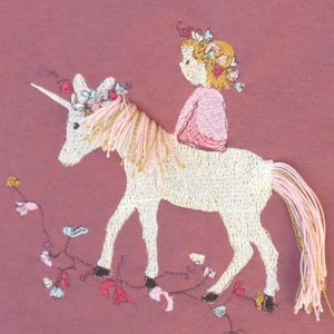May include: Dusty rose-colored fabric with an embroidered design of a girl riding a white unicorn. The unicorn has a pink mane and tail, and the girl wears a pink dress and a floral crown. Delicate floral accents surround the unicorn.