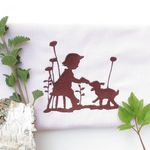 May include: A white fabric with a brown embroidered design of a girl and a lamb. The fabric is surrounded by green leaves and branches.