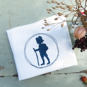 May include: A white tea towel with a blue embroidered design of a person walking with a stick. The tea towel is on a light blue wooden surface with dried flowers and berries.