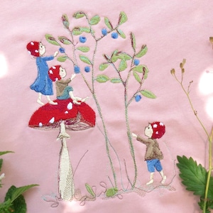 Op de afbeelding: Een lichtroze sweatshirt met een geborduurd ontwerp van drie figuren met rode hoeden en blauwe en bruine kleding die bessen plukken. Een grote rode paddenstoel met witte stippen staat in het midden van het ontwerp.