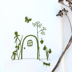 May include: A white tea towel with a green embroidered design of a small house with a door and window, surrounded by flowers and a butterfly. A dried brown plant is in the background.