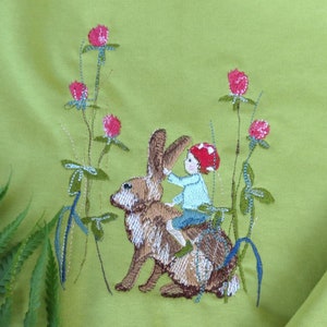 May include: Lime green sweatshirt featuring an embroidered design of a child riding a rabbit through a field of clover. The child wears a red mushroom cap and blue shirt. The rabbit is brown and white. Fern leaves are in the foreground.