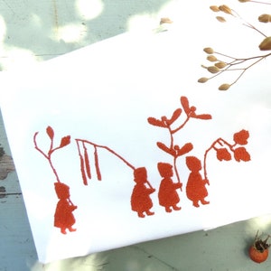 May include: A white tea towel with a red embroidered design of children playing with branches and leaves. The tea towel is lying on a light blue wooden surface with dried flowers and berries.