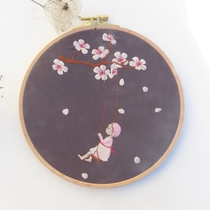 May include: Embroidered hoop art featuring a girl on a swing beneath a blooming cherry blossom tree on a gray background.