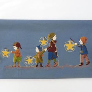 May include: Embroidered textile art showing four children with star-shaped lanterns. The children are in colourful clothes, walking on a light blue background. The artwork has a whimsical, folk art style.