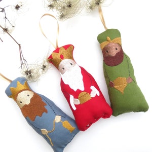 Könnte beinhalten: Drei Filz-Weihnachtsornamente, die die Heiligen Drei Könige darstellen. Jedes Ornament hat eine andere Farbe: Rot, Grün und Blau. Die Ornamente sind mit Details bestickt, darunter Gesichter, Bärte und Kronen.