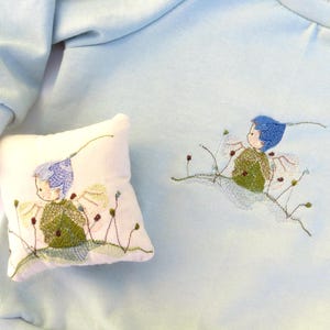 May include: A pale blue sweatshirt and a small pillow, both decorated with embroidered fairy designs. The fairy has blue hair and a green top, with floral details. The pillow is square and features the same fairy design.