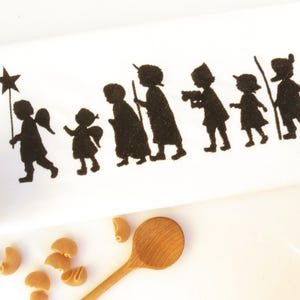 May include: A white fabric with a black silhouette design of a procession of figures, including angels, children, and a shepherd with a sheep. A bowl of pasta and a wooden spoon are in the foreground.