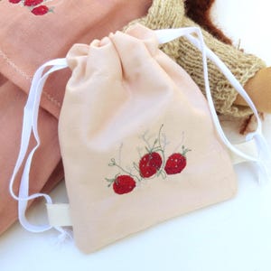 Puede incluir: Una bolsa con cordón de color rosa claro con un diseño bordado a mano de tres fresas rojas. La bolsa tiene cordones blancos y está hecha de una tela suave. La bolsa forma parte de un conjunto con un artículo rosa a juego.