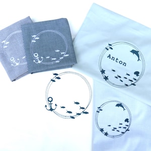 Könnte beinhalten: Mehrere gefaltete Stoffartikel mit nautischen Designs. Zwei quadratische Tücher zeigen einen Anker und Fische in einem kreisförmigen Rahmen. Zwei weiße Tücher tragen den Namen "Anton" und ein Delphindesign. Ein Tuch ist gestreift.