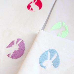 Könnte beinhalten: Weiße Stoffservietten mit gestickten Osterei-Designs. Jede Serviette hat ein andersfarbiges Ei mit einer Kaninchensilhouette im Inneren. Zu den Farben gehören Rosa, Lila, Blau und Grün.