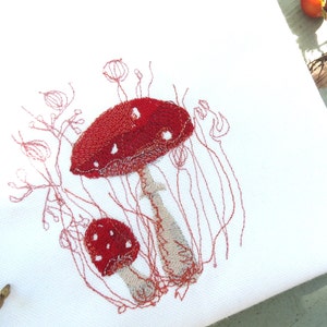 Op de afbeelding: Geborduurd ontwerp van twee rode paddenstoelen met witte stippen op een witte achtergrond. De paddenstoelen zijn omgeven door dunne, roodbruine stelen en bladeren. De borduursels zijn gedetailleerd en op een witte stof.