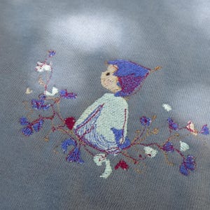 Op de afbeelding: Borduurwerk op een lichtblauwe stof met een klein figuurtje met een paarse hoed en een bloemenmotief.