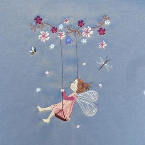 Könnte beinhalten: Stickerei mit einer Fee mit Flügeln, die auf einer Schaukel sitzt, umgeben von rosa und blauen Blumen. Die Fee trägt ein rosa Kleid und hat braunes Haar. Es gibt auch zwei Insekten, eine Biene und eine Libelle, im Design.