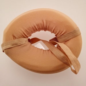 May include: A tan donut-shaped cushion with a beige elastic strap. The cushion is designed to provide support and comfort.