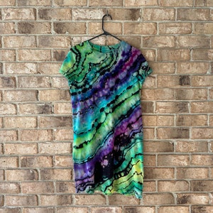May include: A tie-dye dress with a green, purple, and black color scheme. The dress has a loose fit and a round neckline.