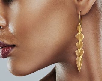 Puede incluir: Una mujer con cabello oscuro lleva un par de aretes largos, dorados, en forma de espiral. Los aretes cuelgan de sus lóbulos de las orejas y están hechos de un metal delgado y retorcido.