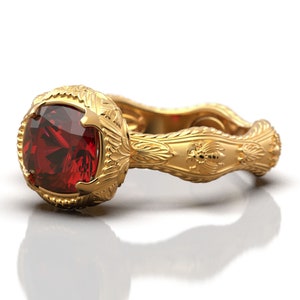 May include: A gold ring with a large, red gemstone set in a detailed, ornate setting. The band of the ring is decorated with intricate carvings and a small bee design.