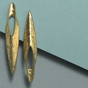 May include: A pair of gold earrings with a hammered texture and an oval cutout. The earrings are long and narrow, with a pointed tip.