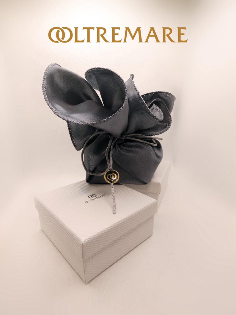 Puede incluir: Una bolsa de terciopelo gris con un logotipo dorado, atada con una cuerda, se encuentra sobre una caja blanca con el logotipo "OLTREMARE" impreso en ella.