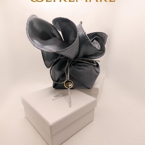 May include: A gray leather pouch tied with a string, sitting atop a white textured box. The pouch is adorned with a gold-colored charm. The box has the word "OLTREMARE" printed on it.