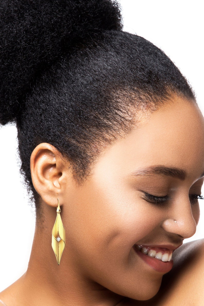 Puede incluir: Una mujer con un peinado afro alto lleva un par de pendientes de oro con un solo diamante en cada pendiente. Los pendientes tienen forma de hoja con un giro.