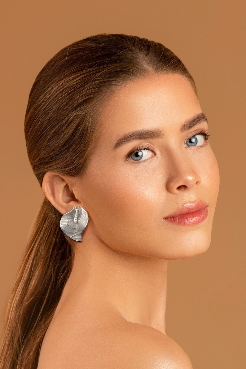 Puede incluir: Una mujer con el pelo largo y casta&ntilde;o y un maquillaje ligero lleva pendientes de plata en forma de hoja. Su piel es lisa y sus ojos son azules.