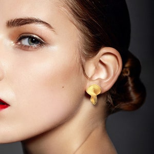 May include: A woman with brown hair wearing gold earrings with a textured, layered design. The earrings are shaped like a teardrop.