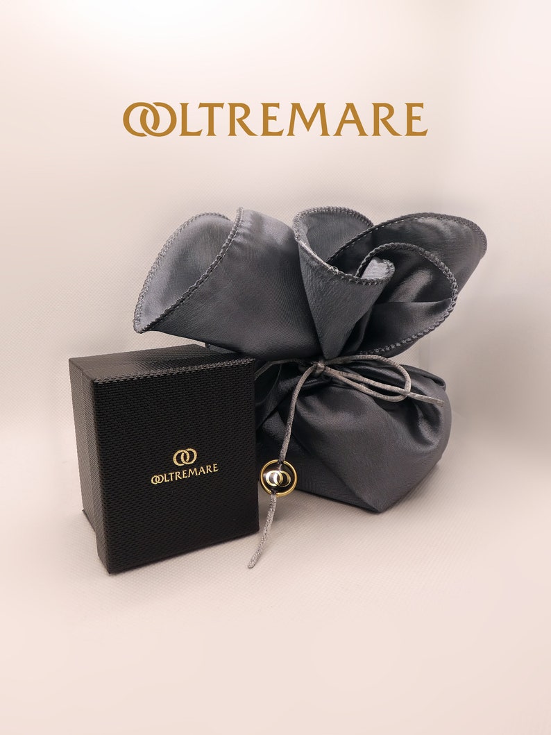 May include: A black gift box with the logo "OLTREMARE" in gold lettering. The box is wrapped in a gray satin pouch with a gold drawstring.