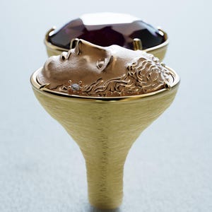 May include: A gold-toned ring featuring a sculpted face with intricate details. The ring has a textured band and a bezel-set, dark purple gemstone. The design is elegant and artistic, suitable for jewelry.