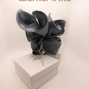 Puede incluir: Una bolsa de cuero gris con forma de flor, atada con una cuerda, sobre una caja blanca. La caja tiene la palabra "OLTREMARE" impresa en dorado. Un dije dorado cuelga de la bolsa.