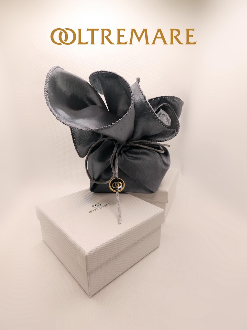 Puede incluir: Una bolsa de cuero gris con forma de flor, atada con una cuerda, sobre una caja blanca. La caja tiene la palabra "OLTREMARE" impresa en dorado. Un dije dorado cuelga de la bolsa.