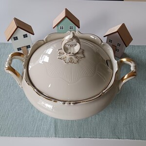 Può includere: Una zuppiera in porcellana bianca con finiture dorate e un coperchio. La zuppiera ha due manici e un pomo decorativo sul coperchio. Tre piccole case di legno sono posizionate intorno alla zuppiera.
