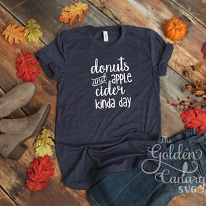 May include: A navy blue t-shirt with the text "donuts and apple cider kinda day" in white lettering. The shirt is laid flat on a wooden surface with fall leaves and a pair of brown boots.