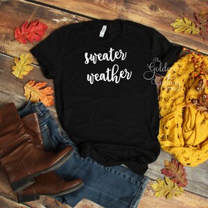 May include: Black t-shirt with the text "sweater weather" in white lettering. The shirt is laying on a wooden surface with jeans, boots, and fall leaves.