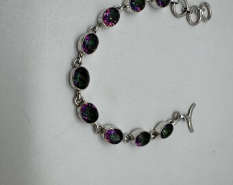 Peut inclure: Un bracelet en argent avec une rangée de pierres précieuses ovales irisées. Les pierres précieuses présentent un mélange de teintes violettes, vertes et noires.