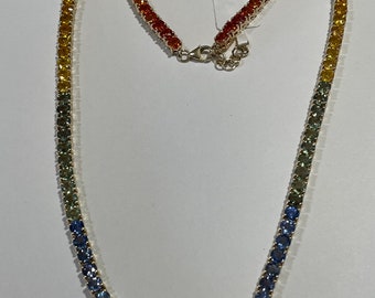 May include: A gold necklace with a gradient of colorful gemstones. The gemstones transition from red to orange, yellow, green, blue, and pink. The necklace has a clasp closure and a tag with text.