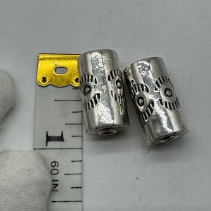 May include: Two cylindrical silver-toned beads featuring an embossed eye pattern, positioned next to a yellow and white measuring tape that indicates each bead is approximately 0.5 inches in length.