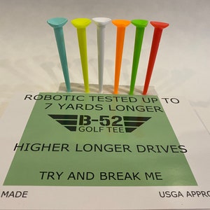 May include: Six golf tees in different colors: light blue, yellow, white, orange, green, and red. The tees are arranged in a row on a white surface. The text "ROBOTIC TESTED UP TO 7 YARDS LONGER" is printed on a green background below the tees. The text "B-52 GOLF TEE" is printed in black below the first line of text. The text "HIGHER LONGER DRIVES" is printed in black below the logo. The text "TRY AND BREAK ME" is printed in black below the second line of text. The text "USA MADE" is printed in black on the left side of the image. The text "USGA APPROVED" is printed in black on the right side of the image.