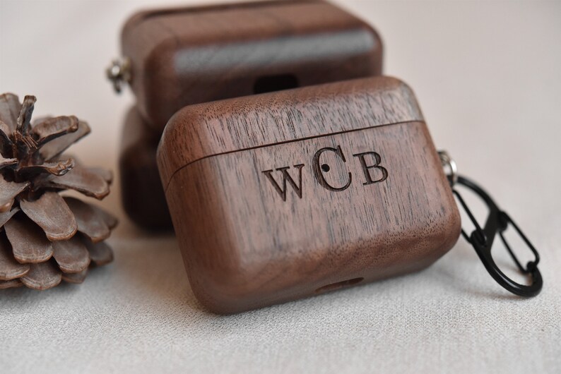 May include: A dark brown wooden case with a carabiner clip and the initials "WCB" engraved on the top. The case is made of walnut wood and has a sleek, modern design.