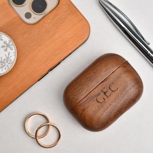 May include: A wooden phone case with a snowflake design phone grip, a wooden Airpods case with the initials "GEC" engraved on it, two gold rings, and a silver pen on a white surface.