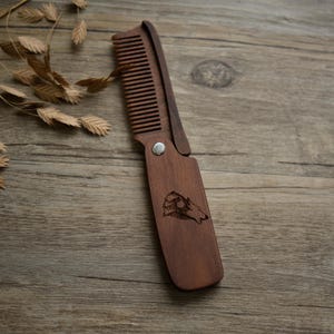 Peut inclure: Un peigne pliant en bois de couleur marron foncé. Le peigne présente une gravure détaillée d'une scène de montagne avec un bélier. Le peigne est ouvert, révélant ses fines dents. Le peigne est en bois et se replie.