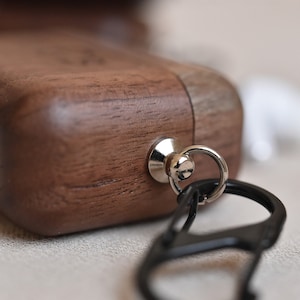 May include: A wooden case with a metal loop and a black carabiner clip. The case is made of dark wood with a natural grain pattern.