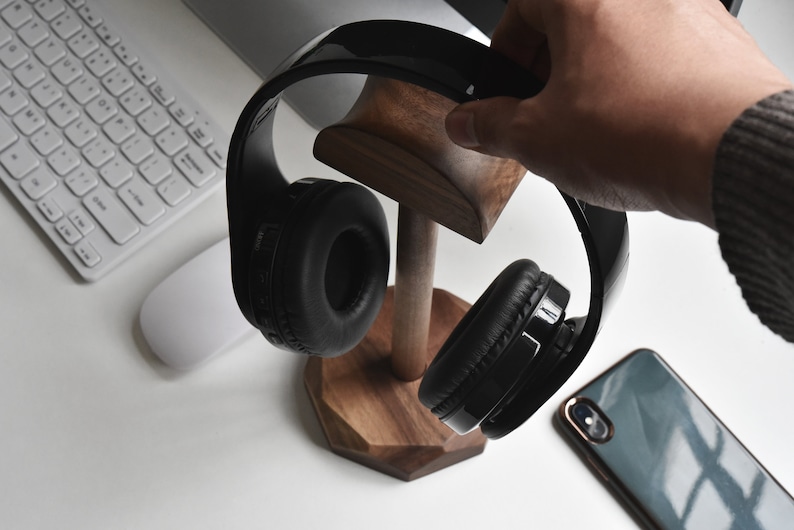 May include: Black headphones with a wooden stand. The headphones are hanging on the stand with the earcups facing out. The stand is made of dark wood and has a hexagonal base.
