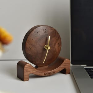 Puede incluir: Un reloj de escritorio de madera marrón oscuro con esfera redonda y manecillas doradas. La esfera tiene el número 12 en la parte superior. La base curva está grabada con el nombre "William".