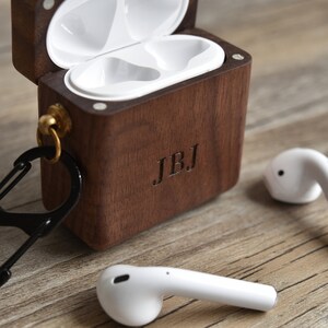 May include: A wooden case with a carabiner clip and the initials "JBJ" engraved on the front. The case is open and two white wireless earbuds are sitting outside of the case.