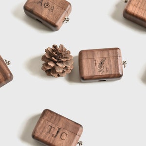 May include: Six wooden AirPod cases with different engravings. The cases are brown and have a small metal loop on the top. One case has the letters "AOA" engraved on it, another has the letters "TJC" engraved on it, another has the letters "a bibby" engraved on it, another has a small bird design engraved on it, and another has a small flower design engraved on it. The cases are arranged in a circle around a small pine cone.