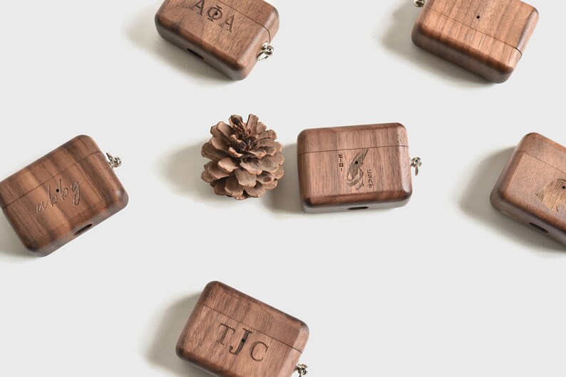 May include: Six wooden AirPod cases with different engravings. The cases are brown and have a small metal loop on the top. One case has the letters "AOA" engraved on it, another has the letters "TJC" engraved on it, another has the letters "a bibby" engraved on it, another has a small bird design engraved on it, and another has a small flower design engraved on it. The cases are arranged in a circle around a small pine cone.